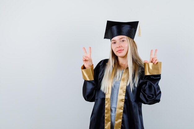 Blondes Mädchen in Abschlusskleid und Mütze, das Friedensgesten zeigt und süß aussieht