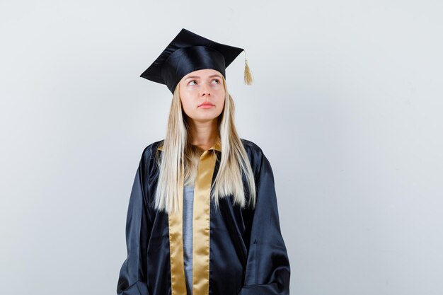 Blondes Mädchen in Abschlusskleid und Mütze, das gerade steht, wegschaut und in die Kamera posiert und nachdenklich aussieht