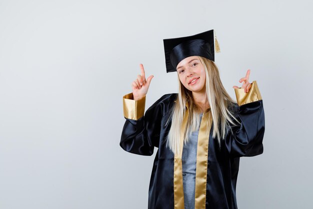 Kostenloses Foto blondes mädchen in abschlusskleid und mütze, das mit den zeigefingern nach oben zeigt und süß aussieht