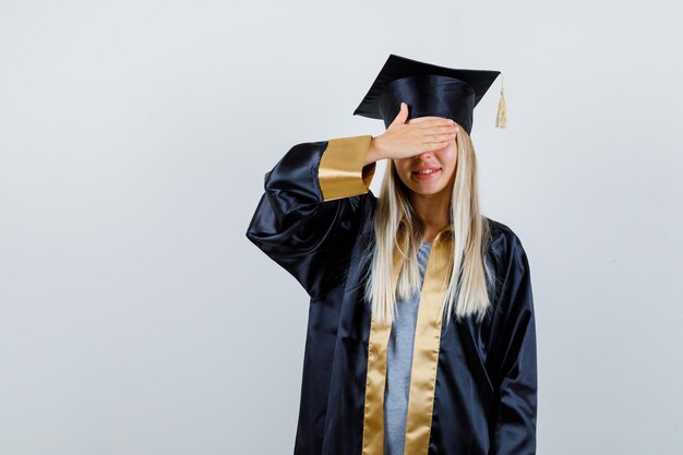 Blondes Mädchen in Abschlusskleid und Mütze, die die Augen bedeckt und glücklich aussieht