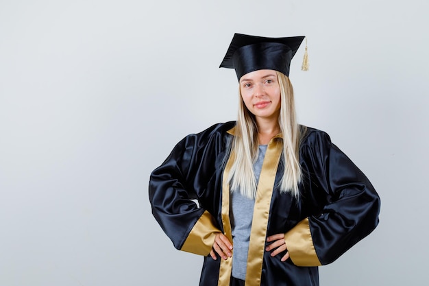 Blondes Mädchen in Abschlusskleid und Mütze, die Hände an der Taille hält und glücklich aussieht