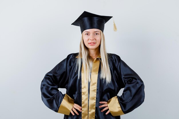 Blondes Mädchen in Abschlusskleid und Mütze, die Hände an der Taille hält und selbstbewusst aussieht