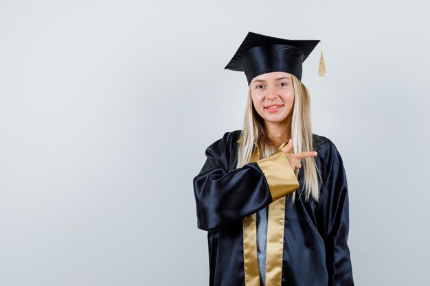 Blondes Mädchen in Abschlusskleid und Mütze, die mit dem Zeigefinger nach rechts zeigt und süß aussieht