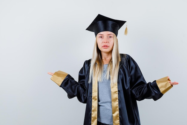 Blondes mädchen in abschlusskleid und mütze, die sich fragend die hände ausstrecken und verwirrt aussehen