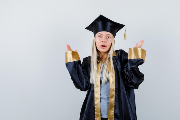 Blondes Mädchen in Absolventenuniform, das hilflose Geste zeigt und verwirrt aussieht