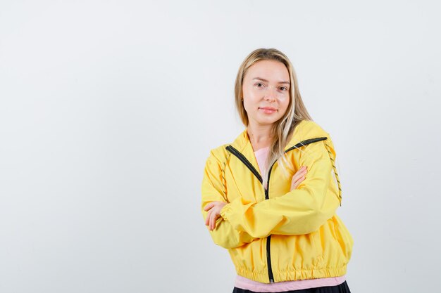 Blondes Mädchen in gelber Jacke, das mit verschränkten Armen steht und hübsch aussieht