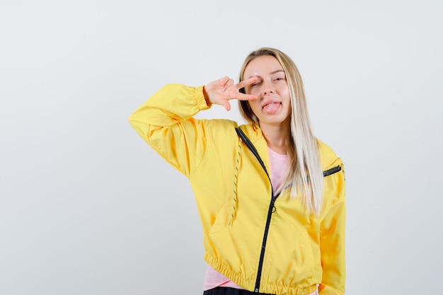 Blondes Mädchen in gelber Jacke, das V-Zeichen in der Nähe des Auges zeigt und glücklich aussieht looking