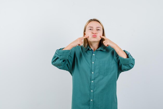 Blondes Mädchen in grüner Bluse, die Wangen aufbläht, mit dem Zeigefinger darauf zeigt und hübsch aussieht