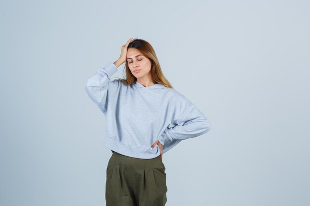 Blondes Mädchen in olivgrünem blauem Sweatshirt und Hose, das eine Hand auf die Stirn legt, während es die Hand an der Taille hält und erschöpft aussieht, Vorderansicht.