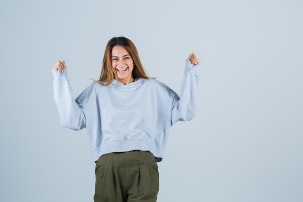 Blondes Mädchen in olivgrünem blauem Sweatshirt und Hosen, die Gewinnergeste zeigen und glücklich aussehen, Vorderansicht.