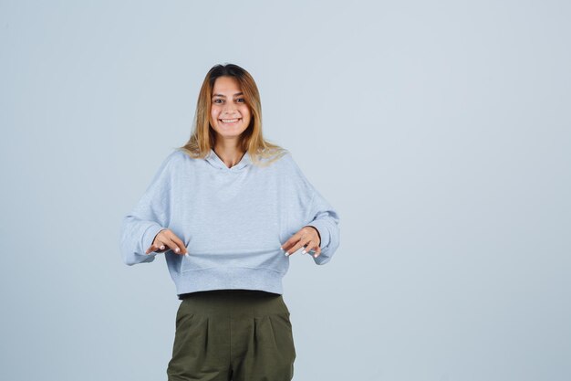 Blondes Mädchen in olivgrünem blauem Sweatshirt und Hosen, die Händchen zum Sweatshirt halten und glücklich aussehen, Vorderansicht.