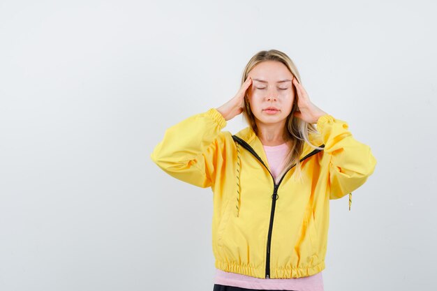 Blondes Mädchen in rosa T-Shirt und gelber Jacke, das die Hände auf die Schläfen legt, die Augen schließt und ruhig aussieht