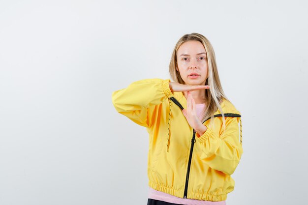 Blondes Mädchen in rosa T-Shirt und gelber Jacke, das eine Zeitpause-Geste zeigt und ernst aussieht