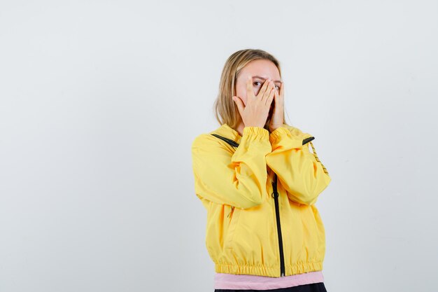 Blondes Mädchen in rosa T-Shirt und gelber Jacke, die das Gesicht mit den Händen bedeckt, durch die Finger schaut und verführerisch aussieht