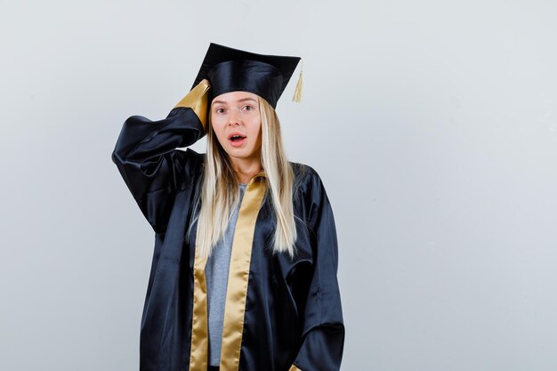 Blondes Mädchen legt Hand auf den Kopf, öffnet den Mund in Abschlusskleid und Mütze und sieht überrascht aus