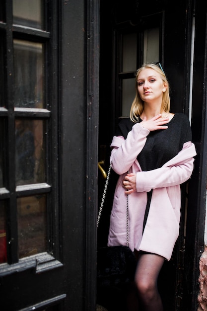 Blondes Mädchen mit Brille und rosa Mantel, schwarze Tunika, die gegen Holztüren gestellt wurde