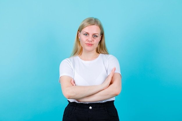 Kostenloses Foto blondes mädchen schaut in die kamera, indem sie die arme auf blauem hintergrund kreuzt