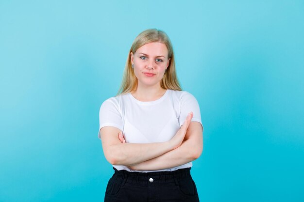 Blondes Mädchen schaut in die Kamera, indem sie die Arme auf blauem Hintergrund kreuzt