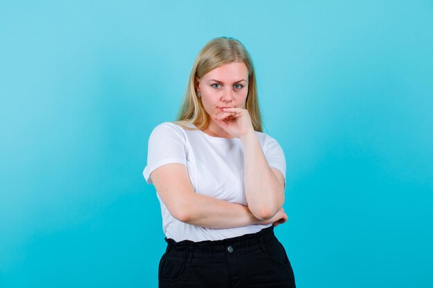 Blondes Mädchen schaut in die Kamera, indem sie die Hand auf das Kinn auf blauem Hintergrund legt