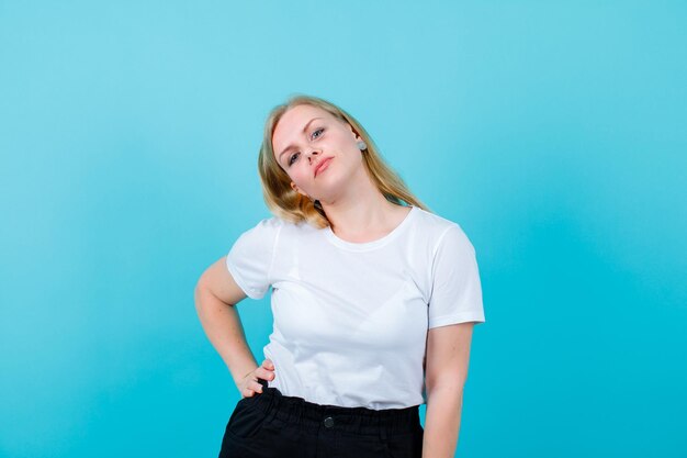 Blondes Mädchen schaut in die Kamera, indem sie die Hand auf die Taille auf blauem Hintergrund legt