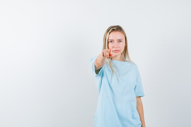 Blondes Mädchen zeigt auf Kamera mit Zeigefinger im blauen T-Shirt und schaut ernst, Vorderansicht.