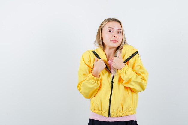 Blondes Mädchen zeigt Daumen nach unten mit beiden Händen in rosa T-Shirt und gelber Jacke und sieht zufrieden aus