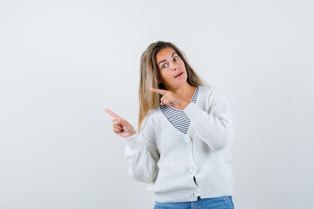 Blondes Mädchen zeigt nach links mit Zeigefingern in gestreiftem T-Shirt, weißer Strickjacke und Jeanshose und sieht verführerisch aus. Vorderansicht.