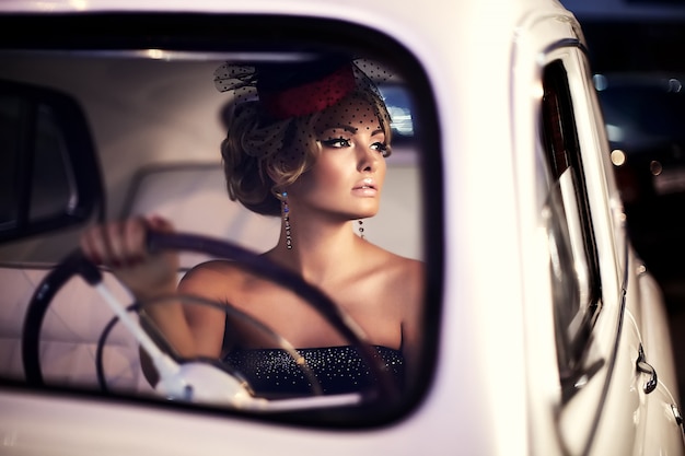 blondes Mädchenmodell der schönen sexy Mode mit hellem Make-up und gelockter Frisur im Retrostil, der im alten Auto sitzt