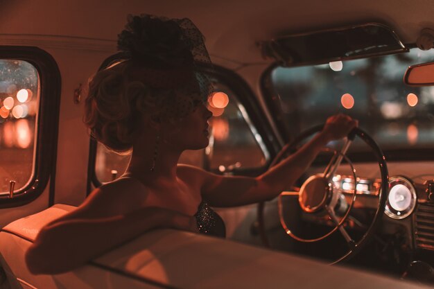 blondes Mädchenmodell der schönen sexy Mode mit hellem Make-up und gelockter Frisur im Retrostil, der im alten Auto sitzt