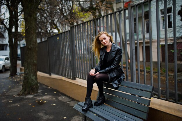 Blondes modisches Mädchen im langen schwarzen Ledermantel posierte auf der Bank