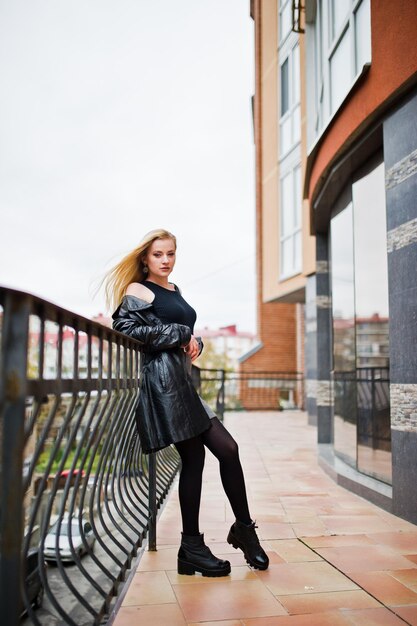 Blondes modisches Mädchen im langen schwarzen Ledermantel posierte vor einem großen Fenster des Gebäudes