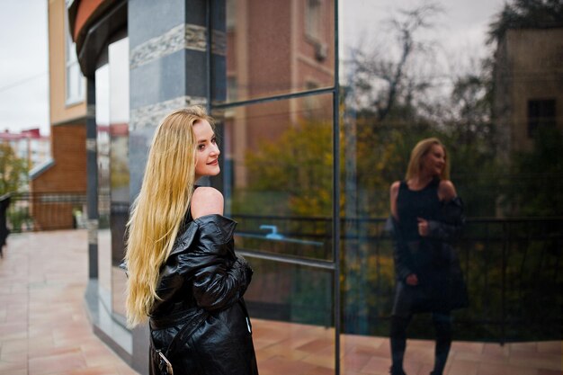 Blondes modisches Mädchen im langen schwarzen Ledermantel posierte vor einem großen Fenster des Gebäudes