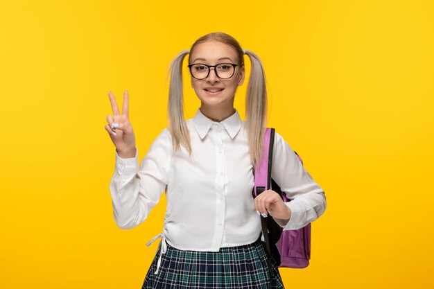 Blondes Schulmädchen des Weltbuchtages, das die Friedensgeste zeigt, die mit Rucksack lächelt