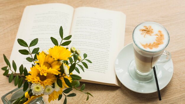 Blühen Sie Vas- und Lattekaffeetasse mit offenem Buch auf hölzerner strukturierter Oberfläche