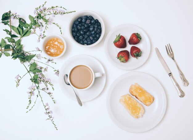 Blume; Marmelade; Blaubeere; Erdbeere; Kaffeetasse und Toastbrot auf weißem Hintergrund mit Besteck