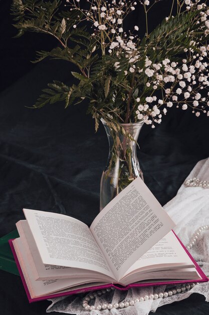 Blumen mit Anlagen im Vase nahe Buch und Perlen auf weißem Gewebe