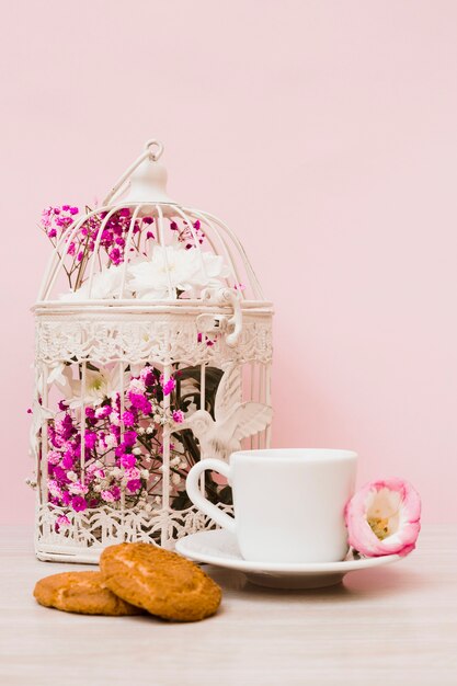 Blumenkäfig; Kaffeetasse und Kekse auf hölzernen Schreibtisch gegen Pastellrosa Hintergrund