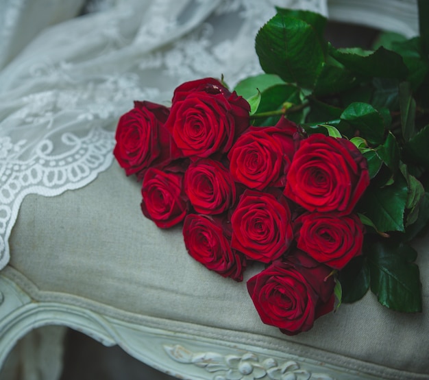 Blumenstrauß der roten Rosen, der auf einem beige Farbstuhl mit Vorhangdetail steht.