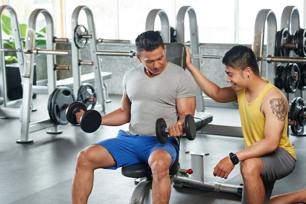 Bodybuilder, der auf anhebenden Dummköpfen der Übungsbank sitzt, die durch persönliche Couch gesteuert werden