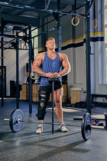 Bodybuilder mit verletztem Bein im Verband hält Langhantel in der Nähe von Cross-Fit-Ständer in einem Fitnessstudio.