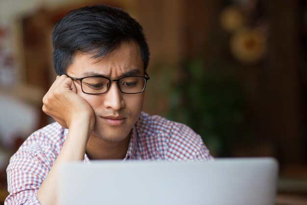 Bored junge asiatische männliche Studentin sitzt am Laptop