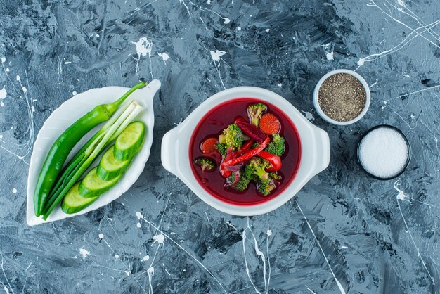 Borschsuppe in einer Schüssel neben frischem Gemüse und Gewürzen auf der blauen Oberfläche