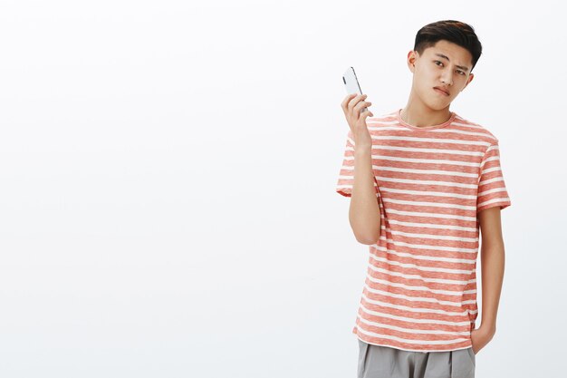 Bossy ernsthaft aussehender cooler junger asiatischer Teenager im gestreiften T-Shirt, der Smartphone als abgelenkt wegnimmt.