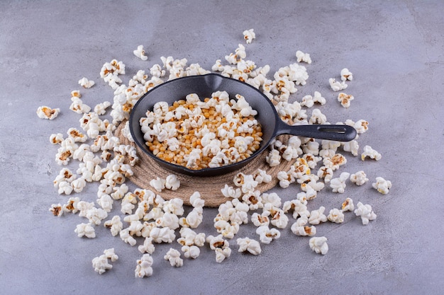 Bratpfanne mit halb aufgeplatzten Maiskörnern, umgeben von einem unordentlichen Popcornkreis auf Marmorhintergrund. Foto in hoher Qualität