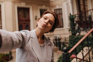 Kostenloses Foto braunäugige dame in grauer jacke nimmt selfie nach draußen und bläst kuss charmante frau in anzug und t-shirt posiert in der nähe eines schönen gebäudes