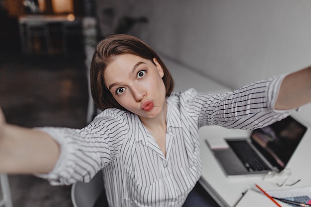 Braunäugige junge Geschäftsfrau macht Selfie mit lustigem Gesichtsausdruck gegen weißen Tisch und Laptop.