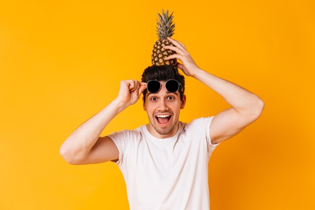 Braunäugiger ungezogener Mann nimmt seine Sonnenbrille ab und hält Ananas auf dem Kopf.