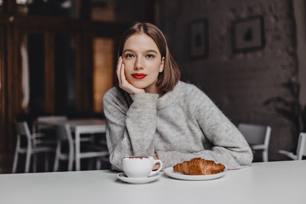 Braunäugiges Mädchen im Wollpullover lehnte sich auf weißen Tisch im Café und schaute in die Kamera. Foto der Frau mit den roten Lippen, die Kaffee und Croissant bestellen.