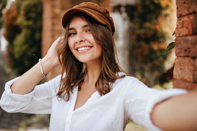 Braunäugiges Mädchen in Samtmütze und weißer Bluse macht Selfie auf Raum von Backsteinmauer und Bäumen.