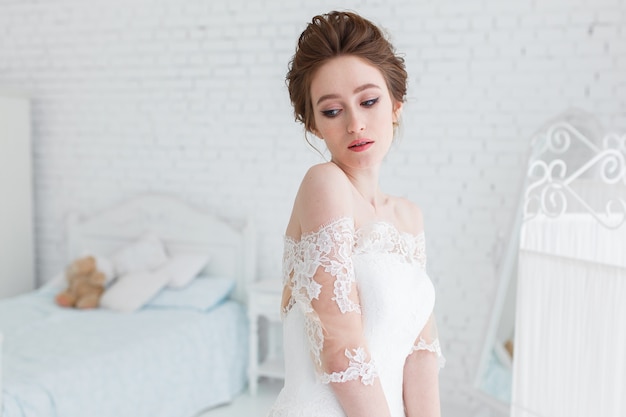 Braut, die im Hochzeitskleid im Studio auf einem Hintergrund einer Backsteinmauer und des weißen Betts aufwirft